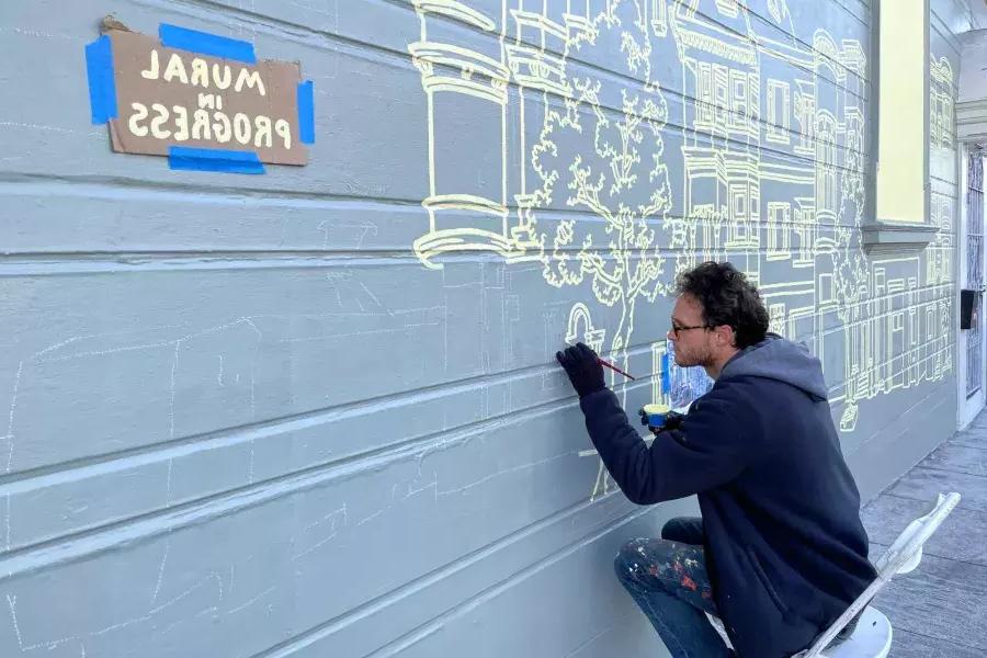 在mission区，一名艺术家在一座建筑的旁边画壁画, 大楼上贴着一个牌子，上面写着“壁画正在进行中”. San Francisco, CA.